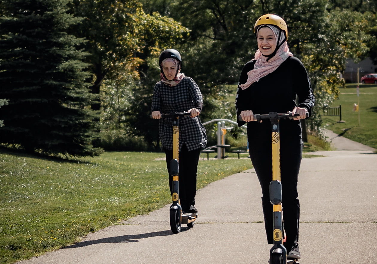 Two people riding Scooty scooters on park path, smiling